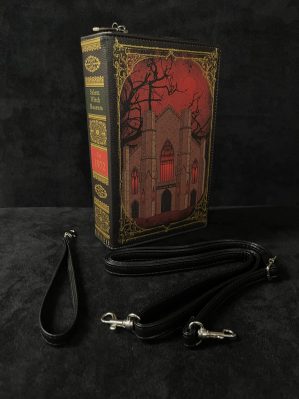 This image features the book purse at a 45 degree angle, showing off the side of the purse with the museum facade against a red backdrop. The "binding" of the book purse is visible, with the words "Salem Witch Museum Est. 1972." Also displayed are the two straps, one for the hand and one for over the shoulder, that come with the purse.