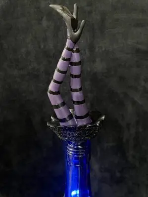 The purple witch legs wine bottle stopper is shown in use. The legs are wearing purple horizontally striped tights with black high heels. Close to the base of the wine stopper, the legs are wearing a sparkly black skirt.