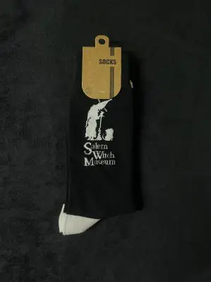 The socks are black, with white heels and toes. The Salem Witch Museum logo, in white, is on the upper portion of the sock.