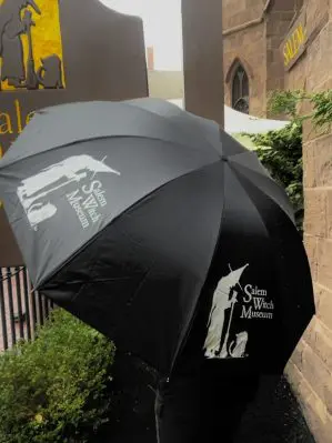 Image shows the umbrella in use. On two out of the umbrella's ten sections, the words "Salem Witch Museum" and our logo, a witch and cat, are emblazoned.