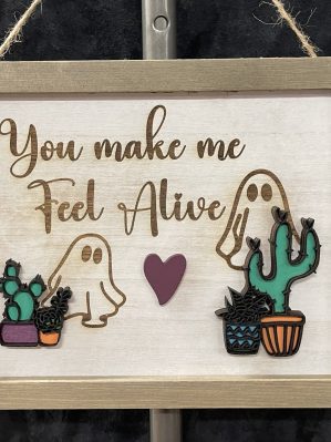 Wooden wall sign hung with twine, featuring two ghosts with their collection of cacti. There is a heart in the center, and the phrase "You make me feel alive" is engraved.