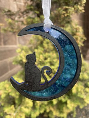 This ornament features an opaque black cat resting in the crescent of a moon with a black outline. A translucent, sparkly, deep azure blue acrylic backing turns the moon part of the ornament into a suncatcher.