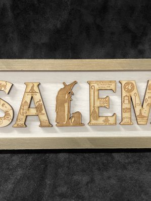 Tan woodcut on a white background spelling out "Salem," with the witch museum logo as the "L." Engraved into the other letters are miniature depictions of iconic Salem scenes. The sign is in a tan frame.